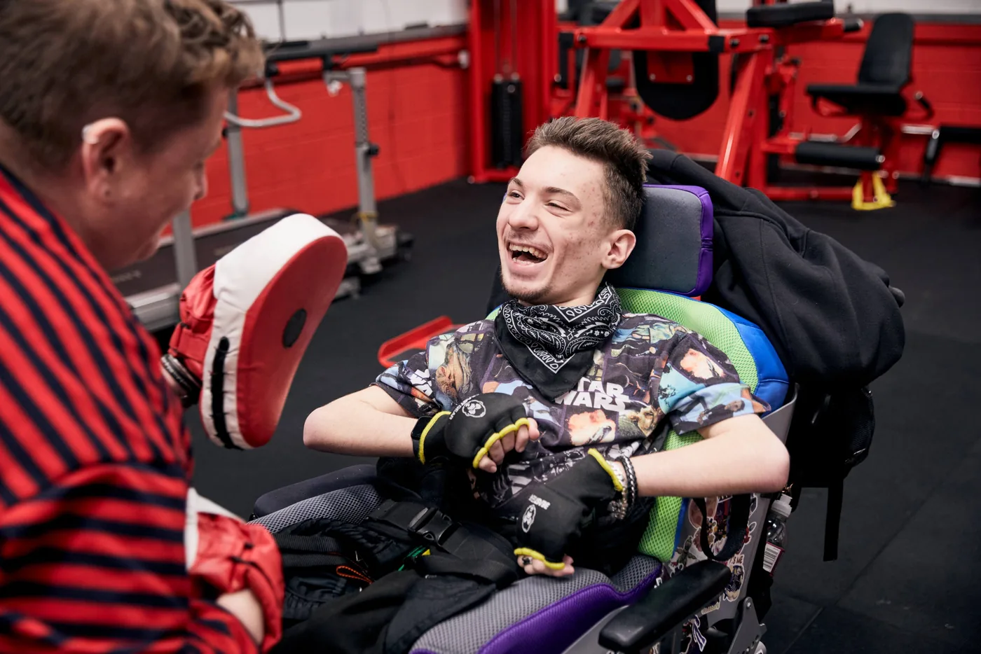 Tyler laughing in his wheelchair at a gym, facing a coach holding a boxing focus pad